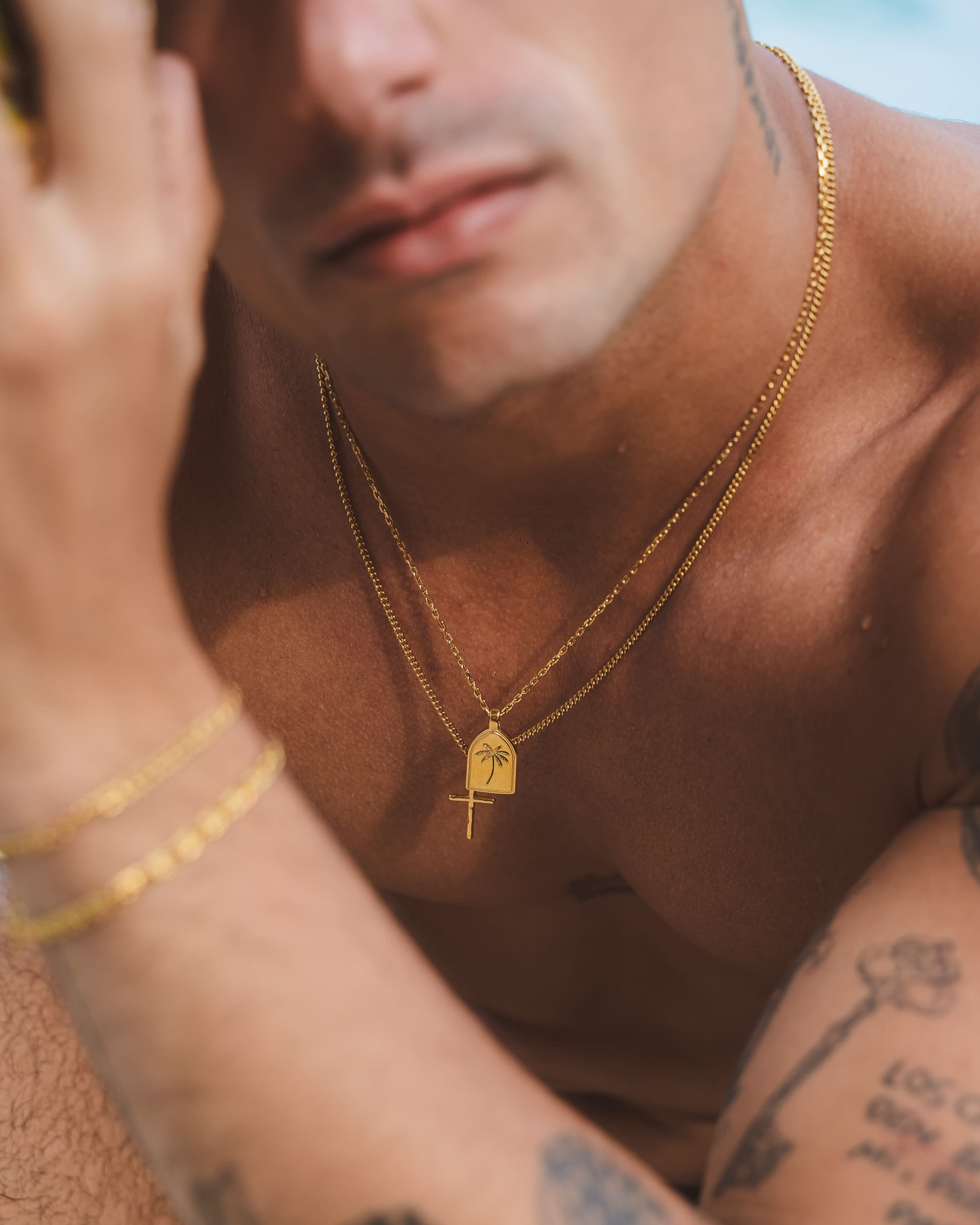Close up of a bearded man wearing a silver chain necklace with a pendant on a tan shirt.