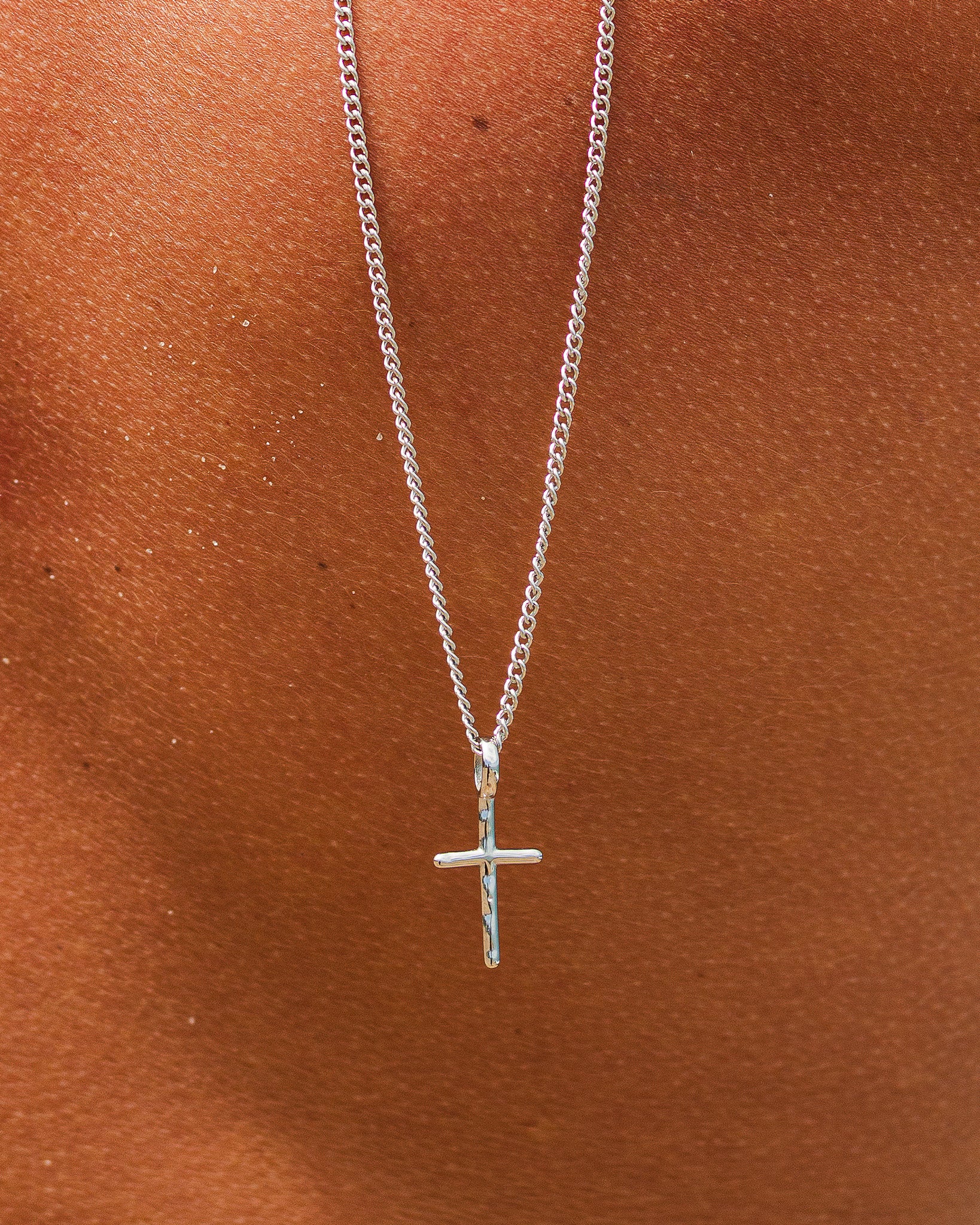 Two silver chain necklaces with a small cross pendant visible at the bottom against a warm brown background.