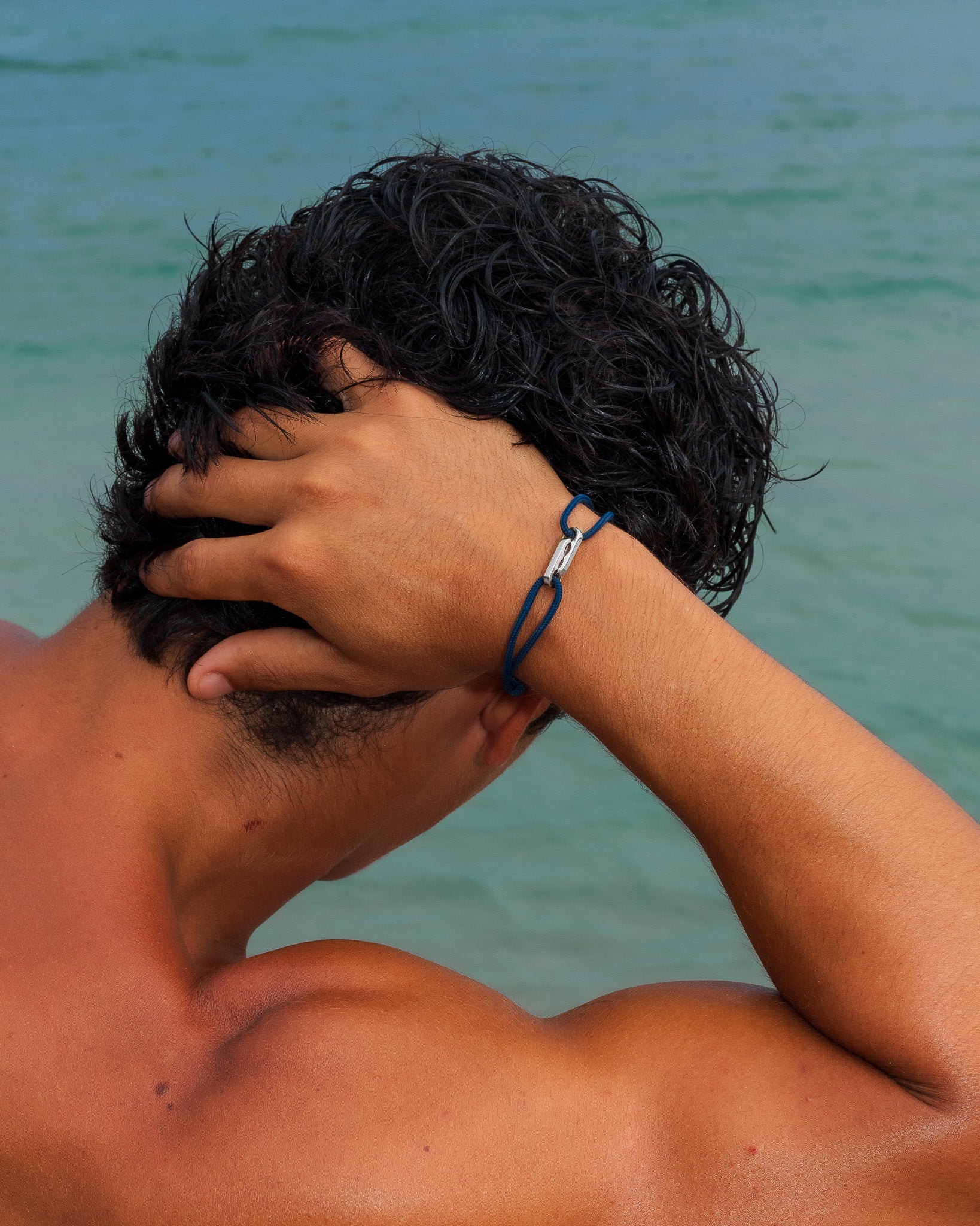Back view of a shirtless man with a blue cord bracelet and silver clasp on his wrist, sea in the background.