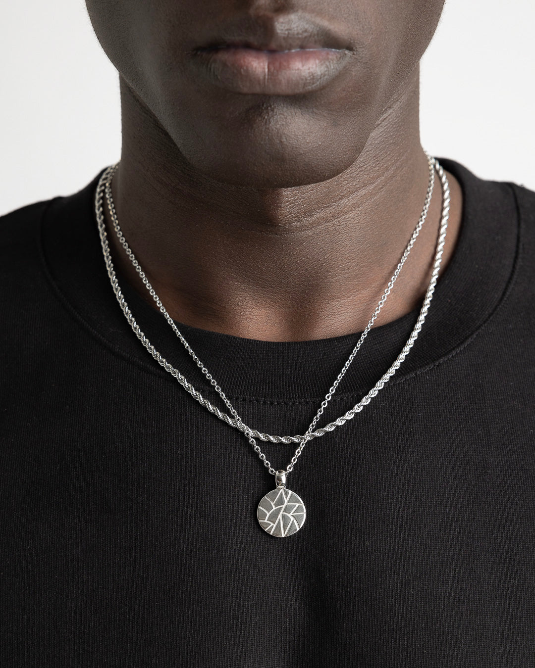 Two silver chains with a round geometric pendant on a model's chest against a black shirt.