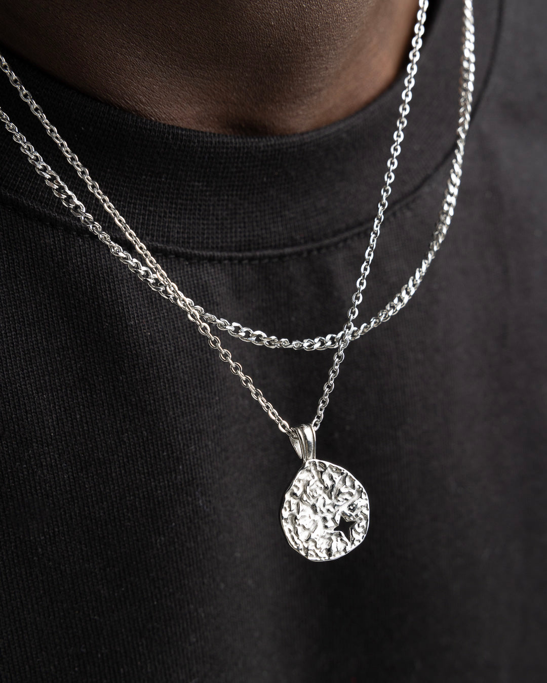 Close up of two silver chains with a textured round pendant resting on a black top.