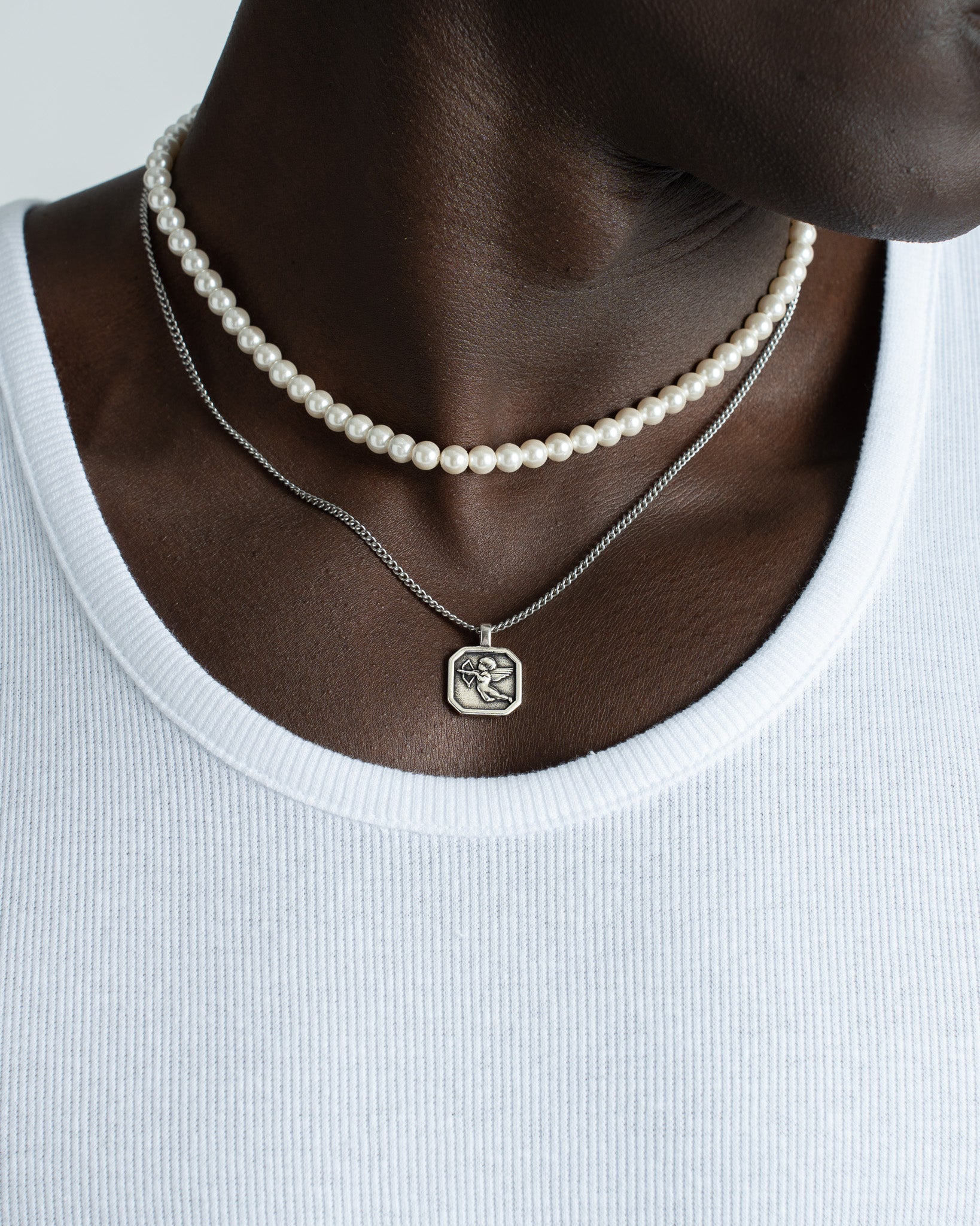 Closeup of neck with a pearl choker and a silver chain necklace featuring Cupid pendant