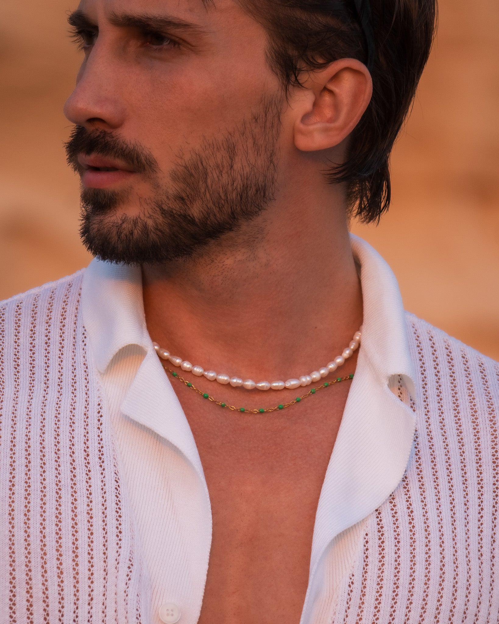Close-up of a man wearing a layered pearl necklace and a gold chain with green beads