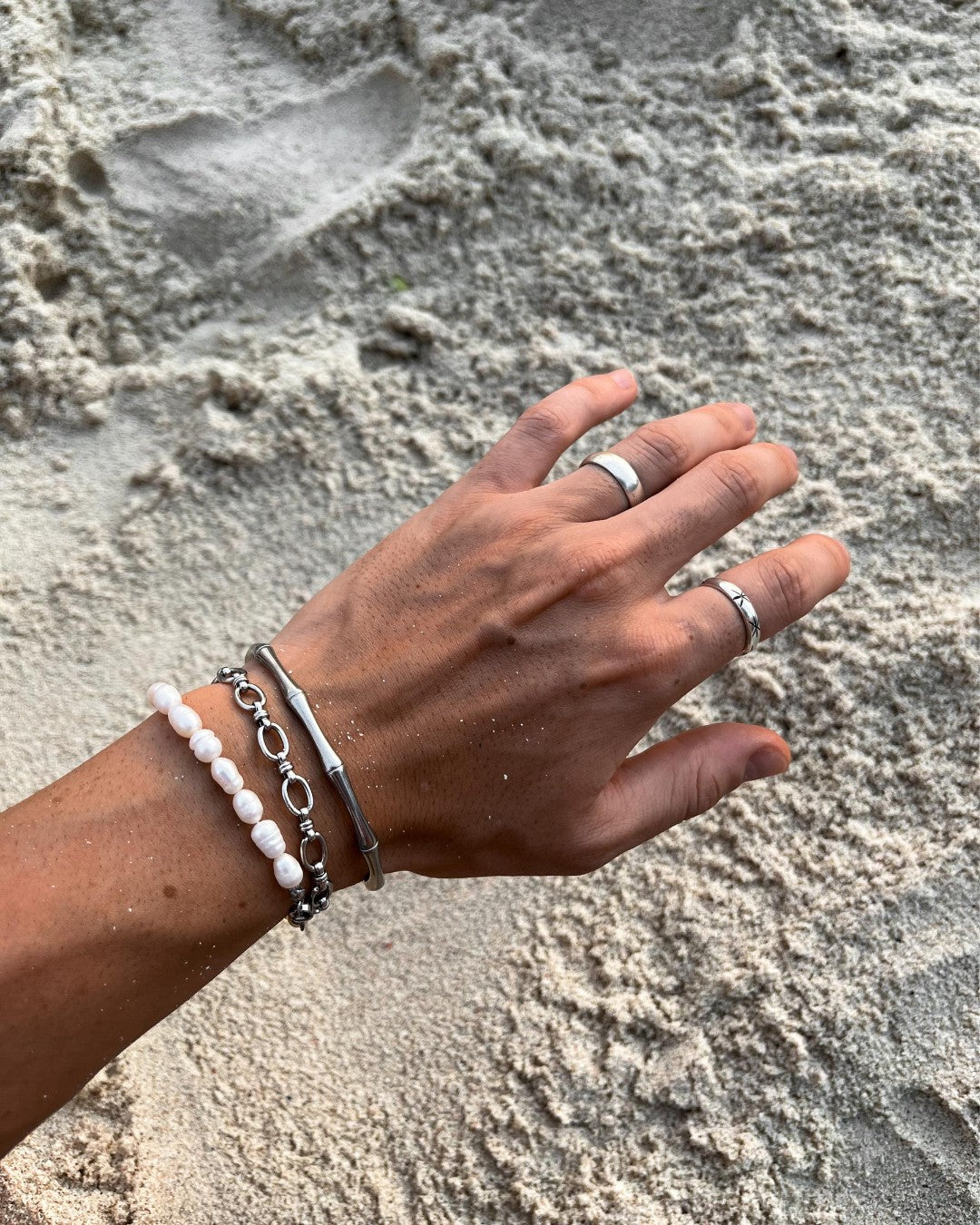Hand wearing bamboo silver bracelet and a white beaded bracelet on a sandy surface