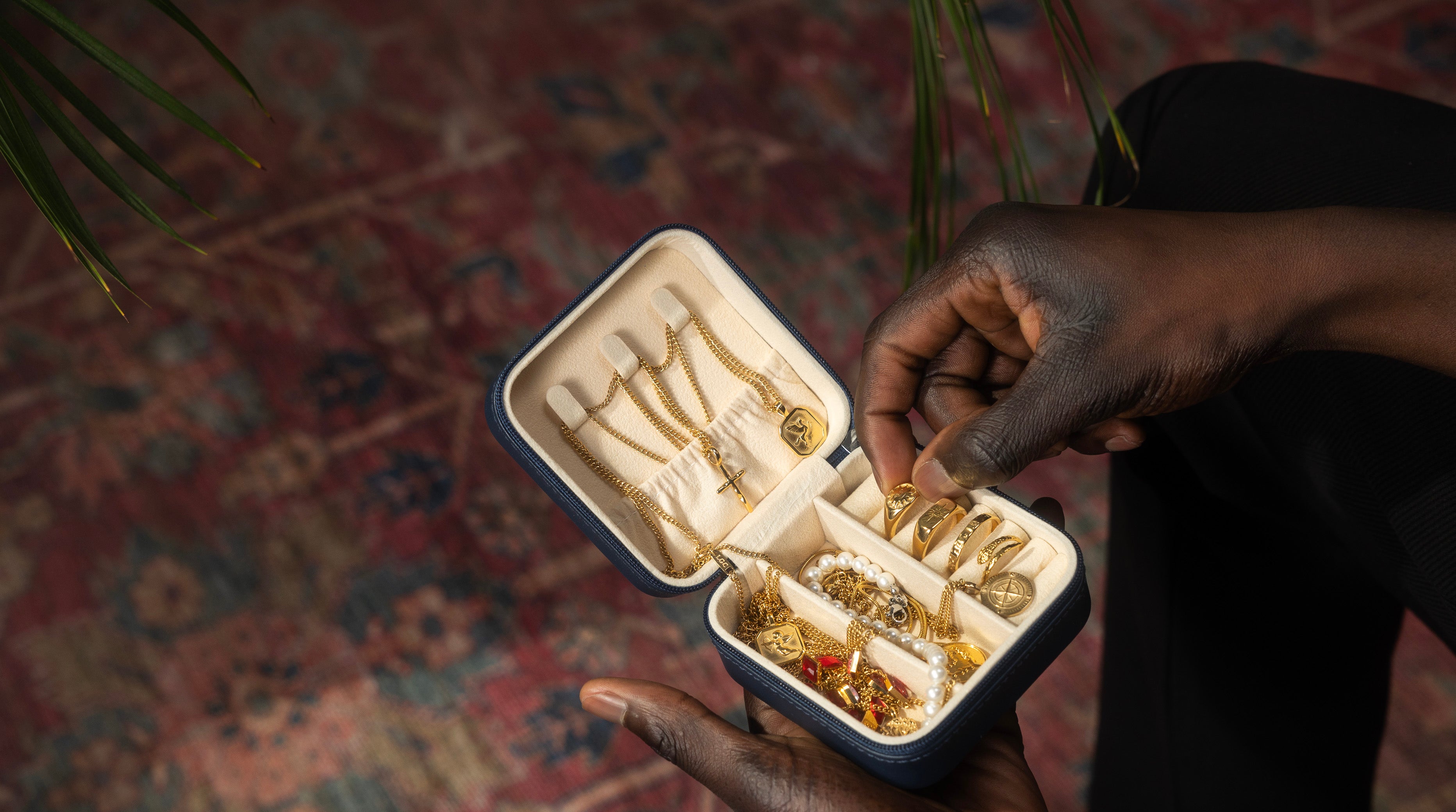 Hands holding an open Lost Gen Club travel jewelry case filled with gold chains, signet rings, pendants and pearls, over a vintage Persian rug.