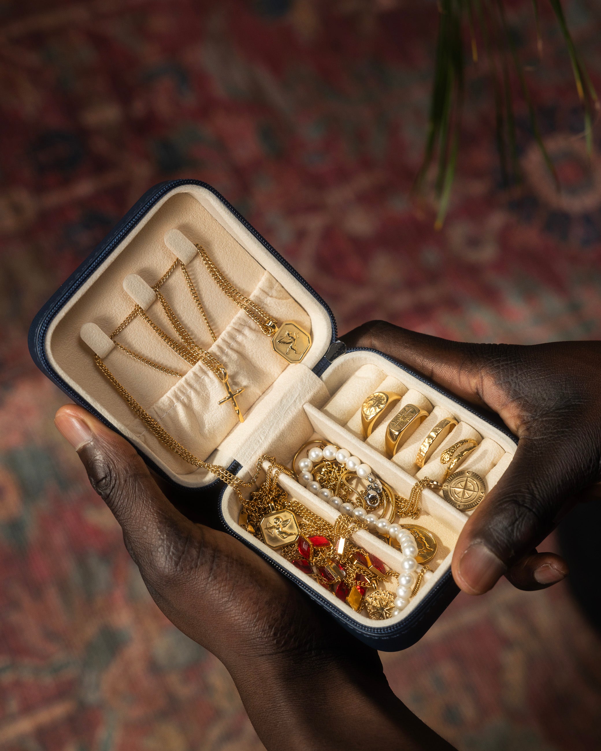 Open blue jewelry case held by dark skinned hands displaying gold chains rings and pearl bracelets inside
