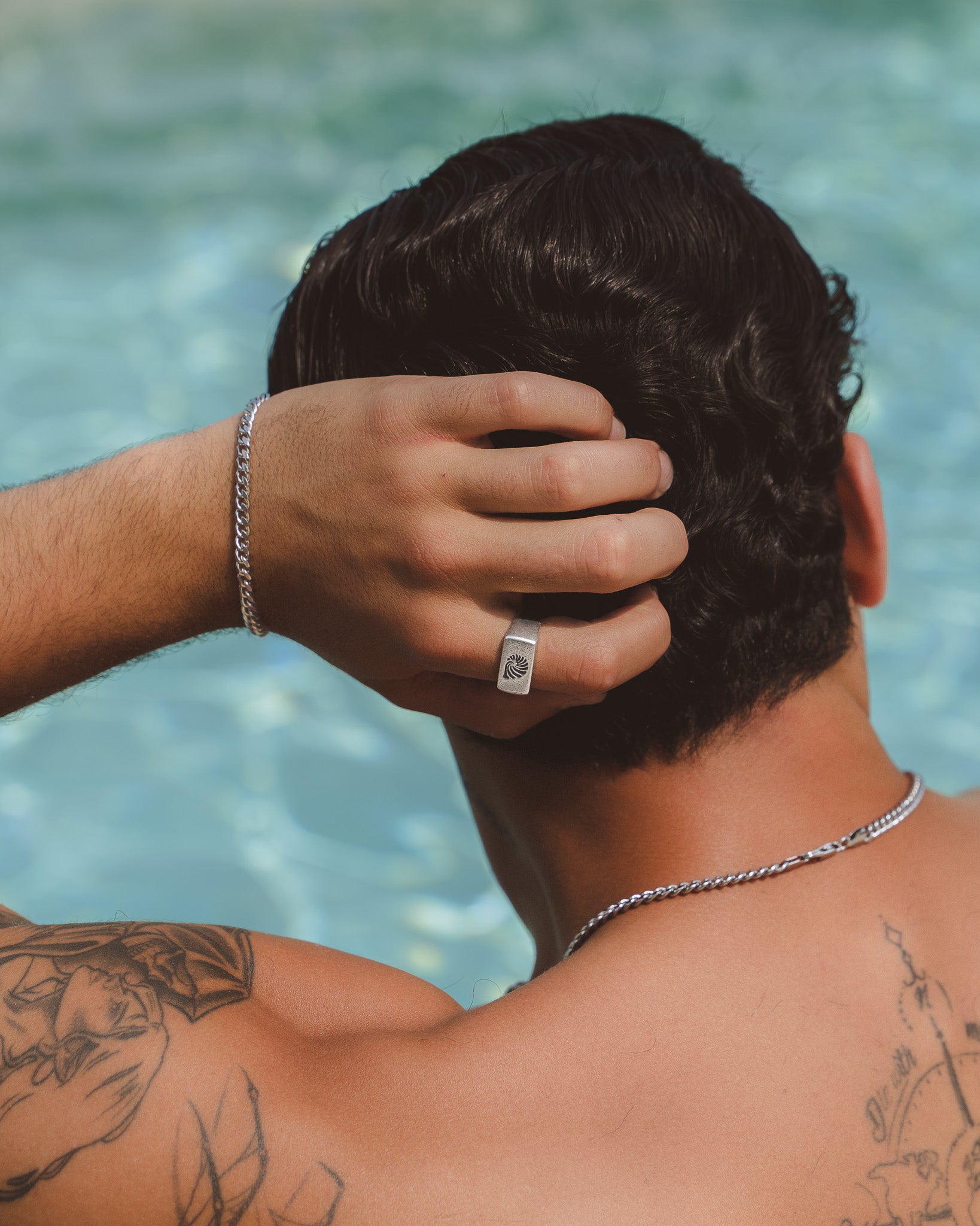 Side view of a man by a pool with a square silver shell ring on his finger and a silver bracelet.