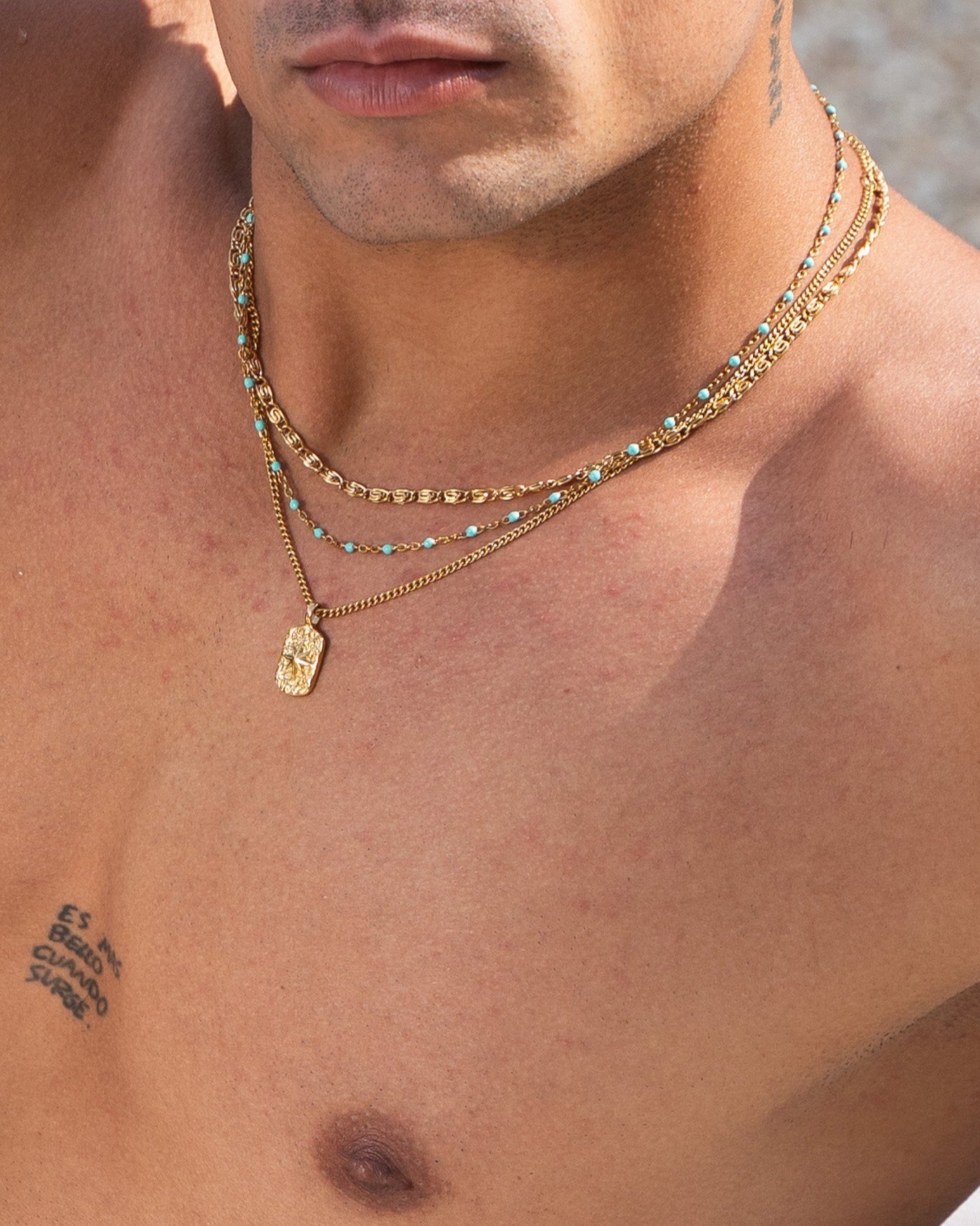 Close up of layered gold chains with turquoise beads and ornate pendants on a man wearing a black shirt