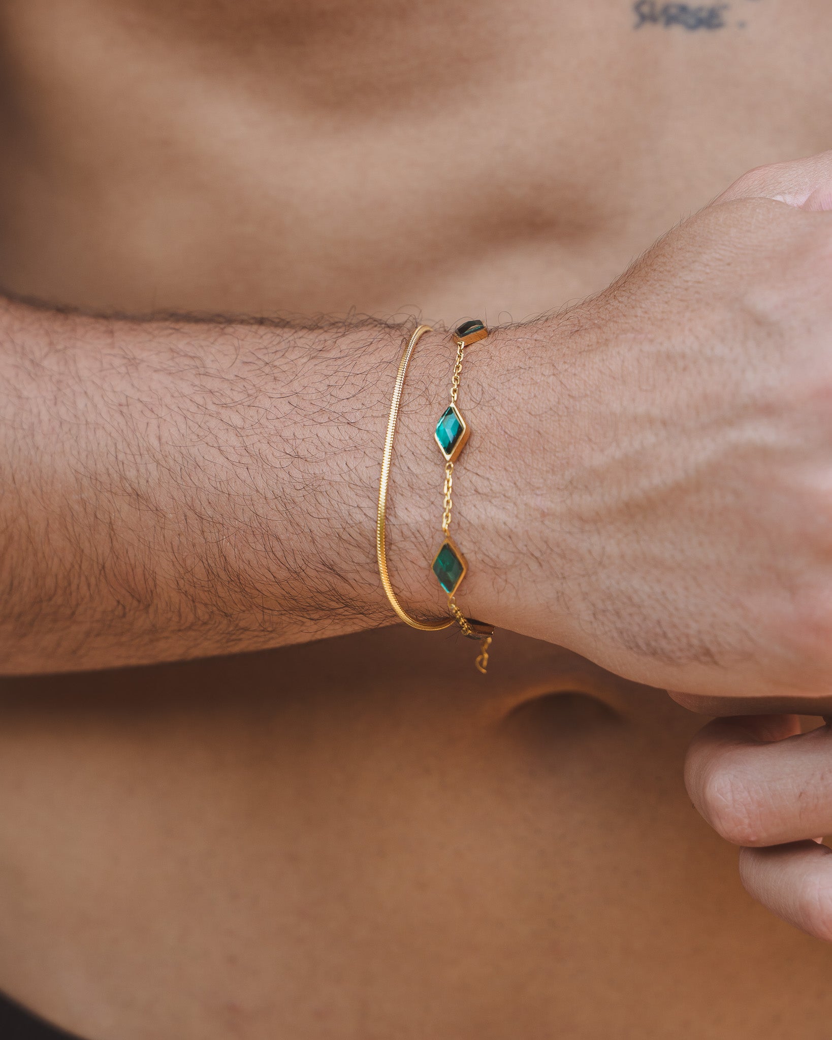 Amberes gold bracelet with emerald green diamond shaped stones on a wrist, paired with a slim gold bracelet.