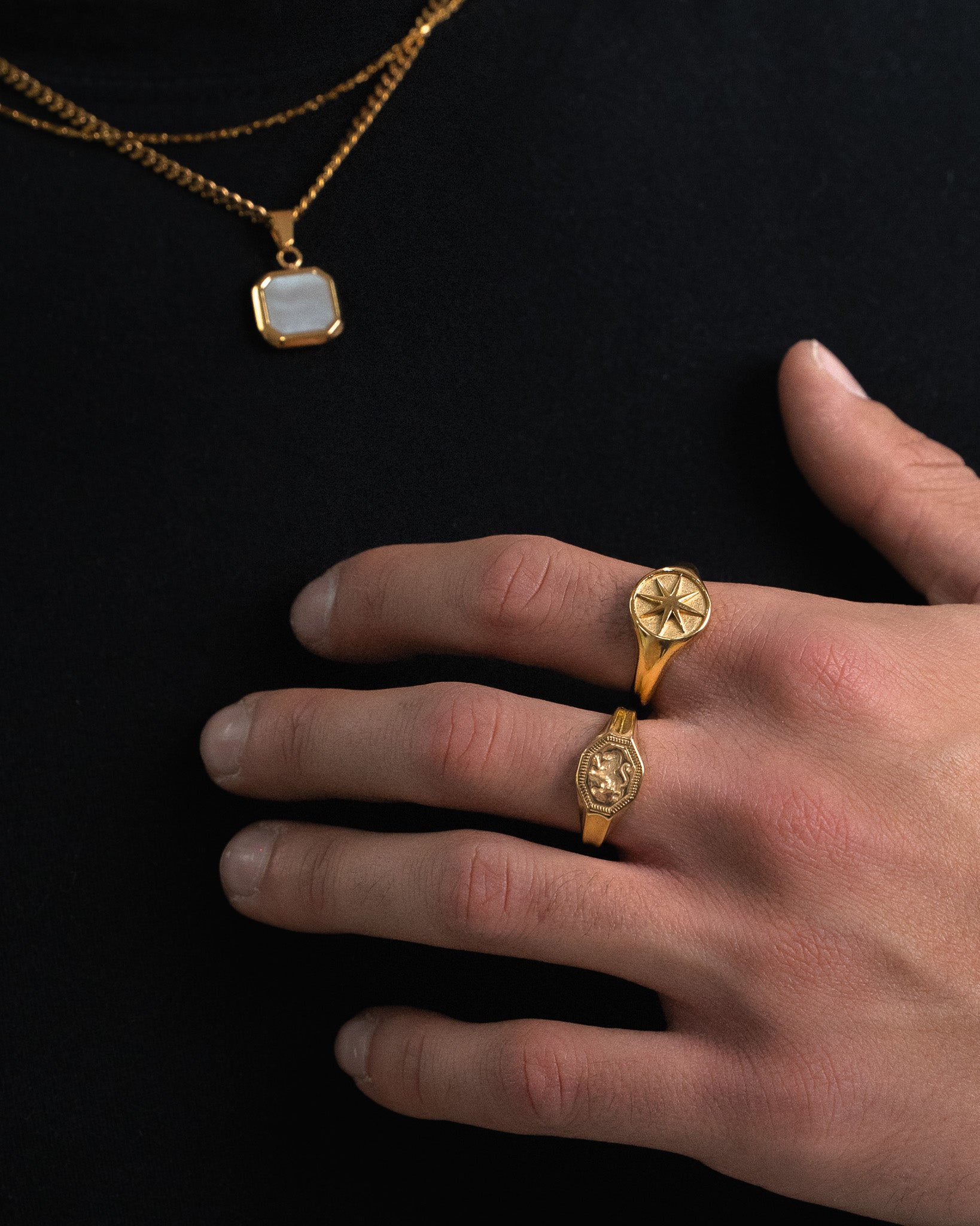 Legacy gold ring with a sunburst emblem worn on the finger beside a gold chain necklace with a square white stone pendant on black background