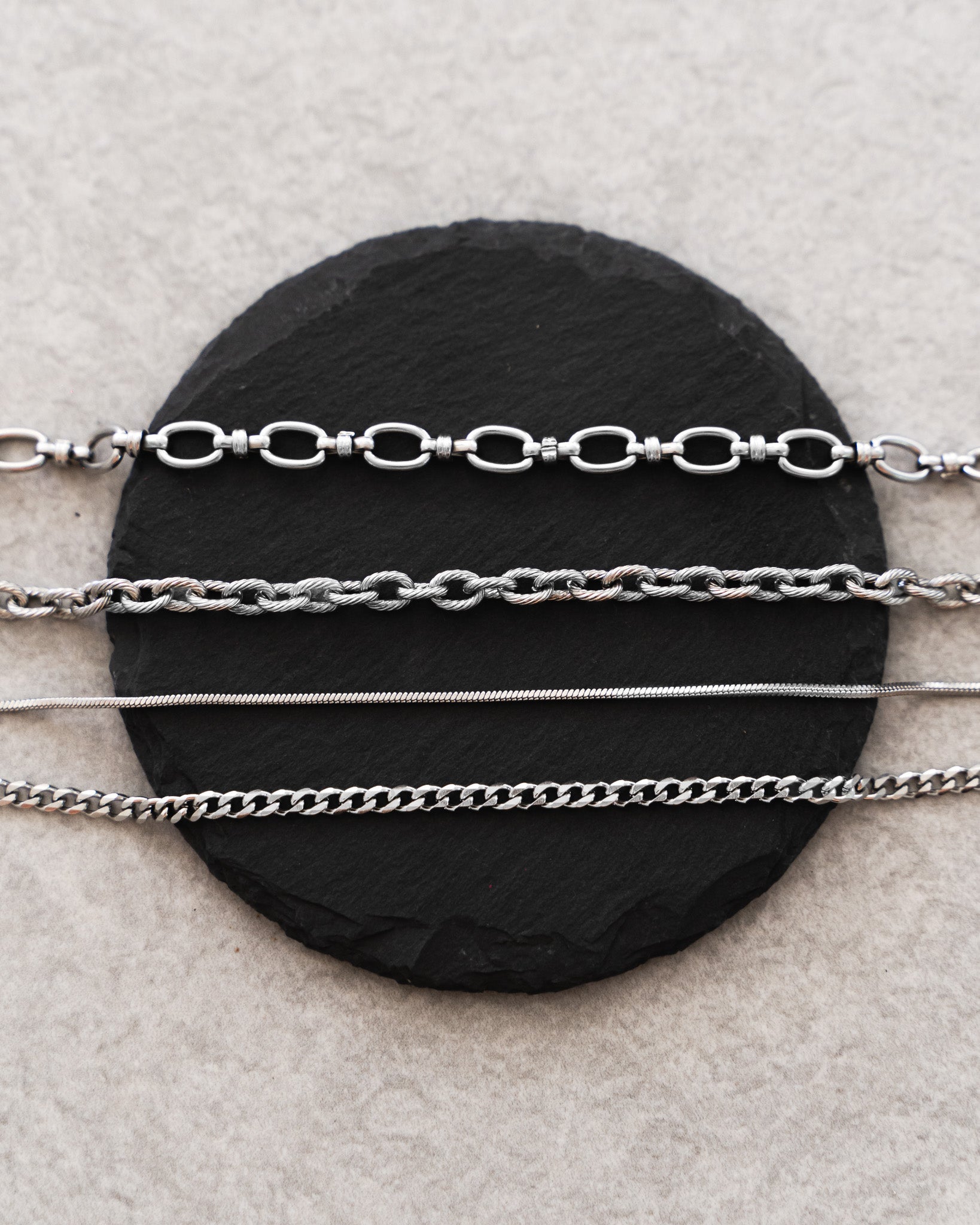 Four silver color bracelets in different link styles displayed on a black slate disc.