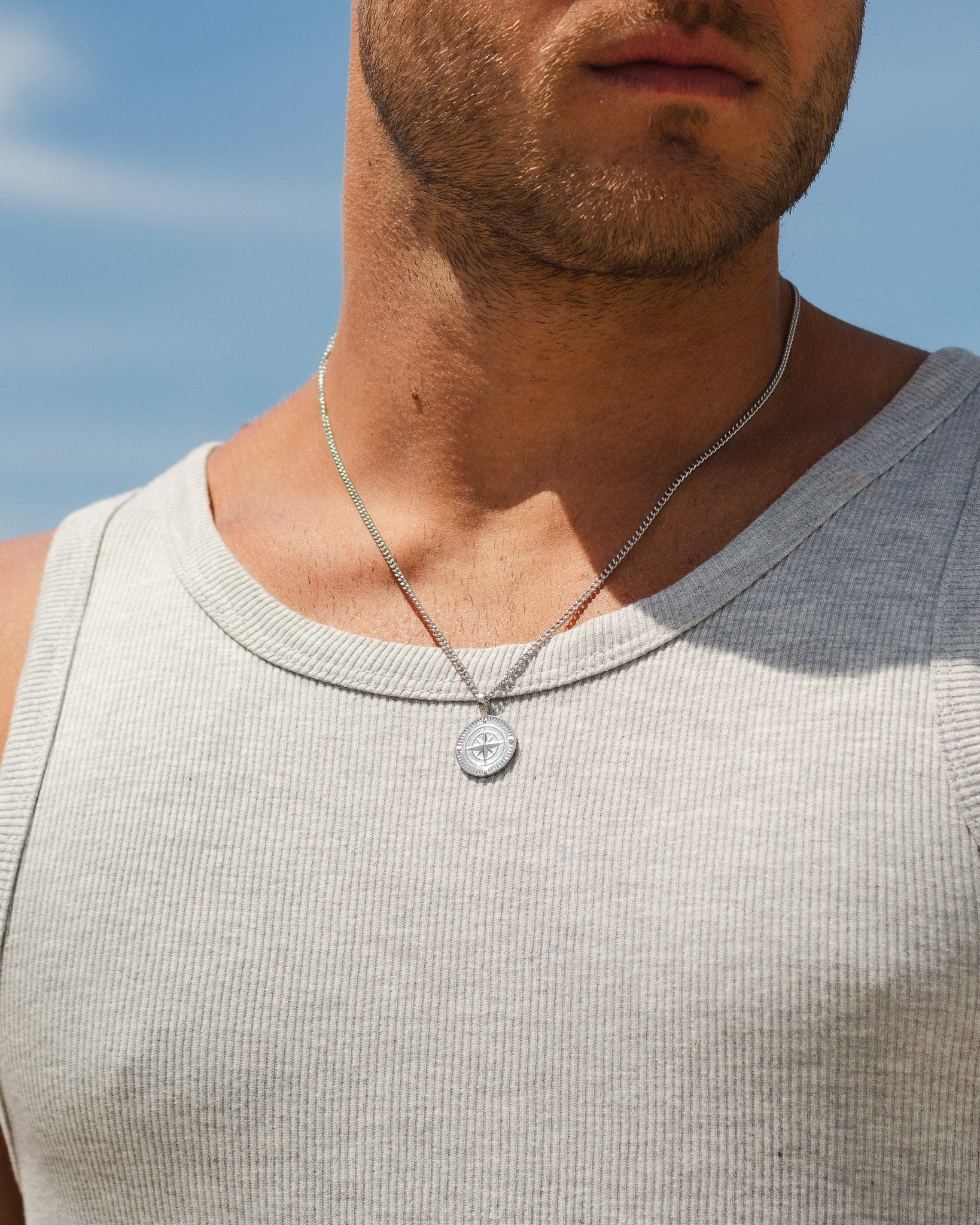 Close-up of a silver compass pendant necklace resting on a man's chest against a blue sky