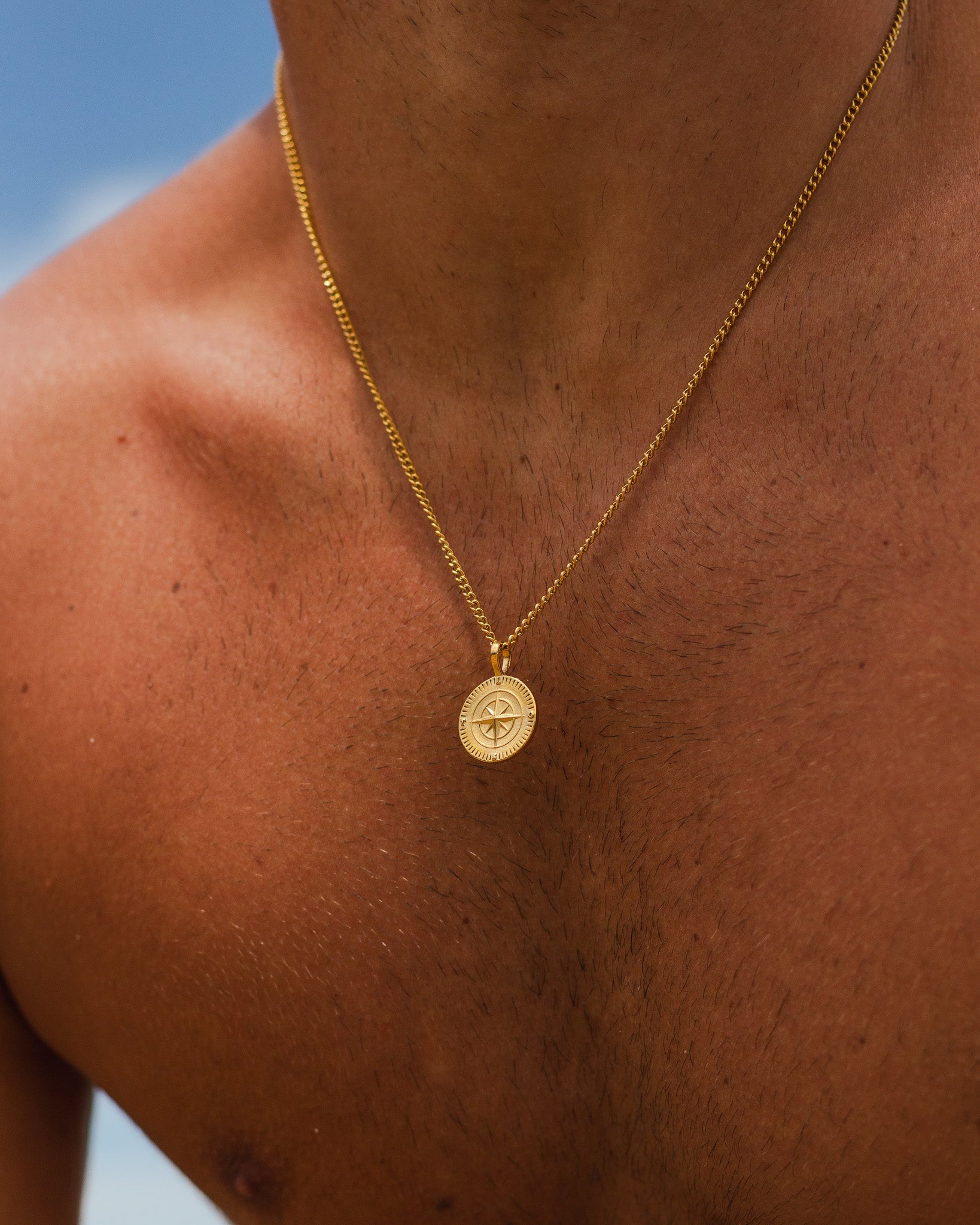 Side view of a man wearing a gold chain necklace with a compass pendant against a blue sky