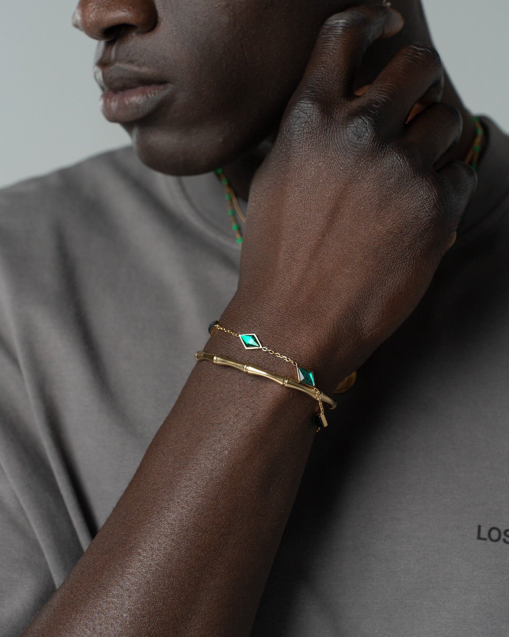 Gold bracelet with emerald-cut green gemstone on wrist, paired with a green beaded bracelet.