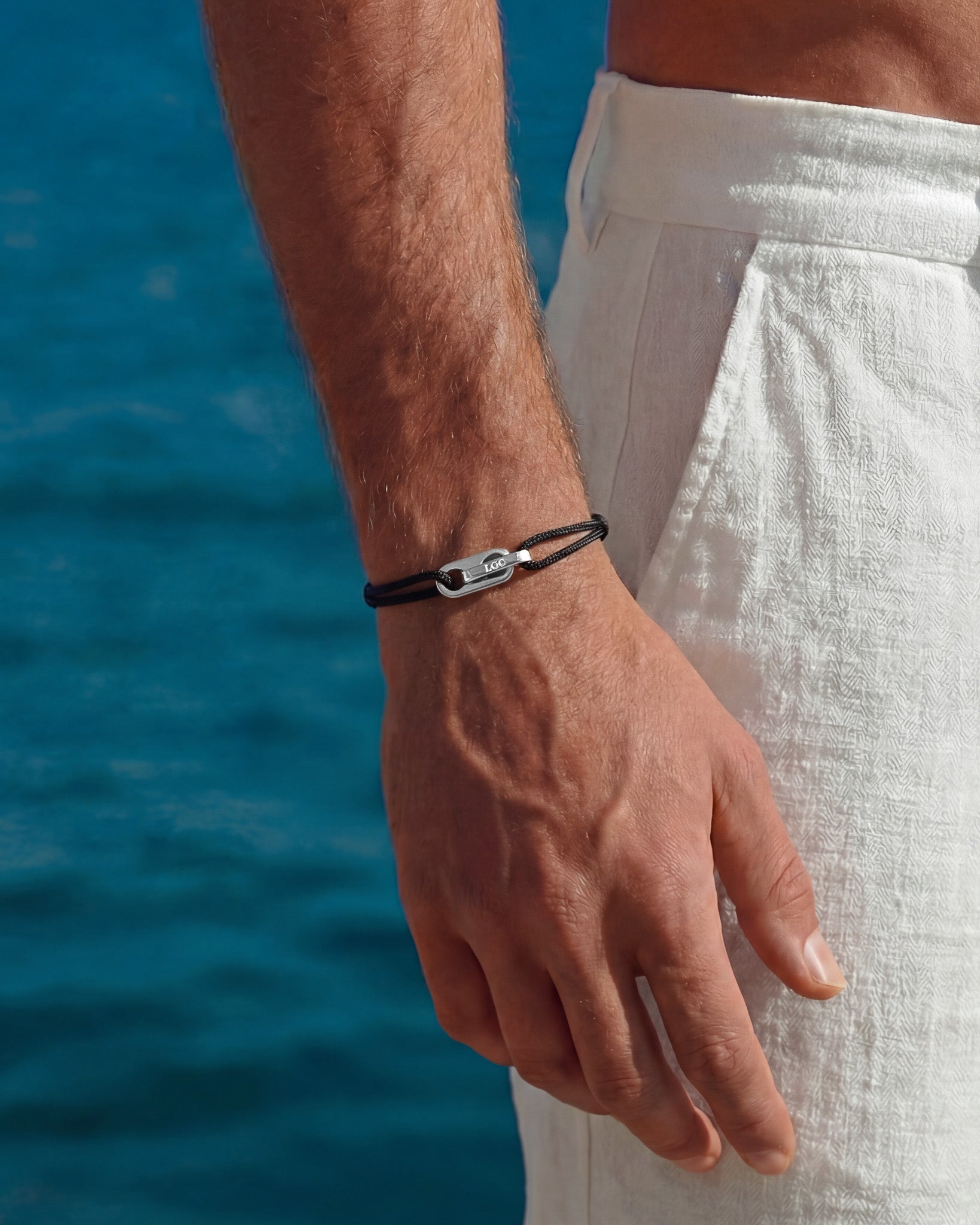 Man wearing an Atlantis silver bracelet with black cord and a silver clasp against a blue ocean backdrop