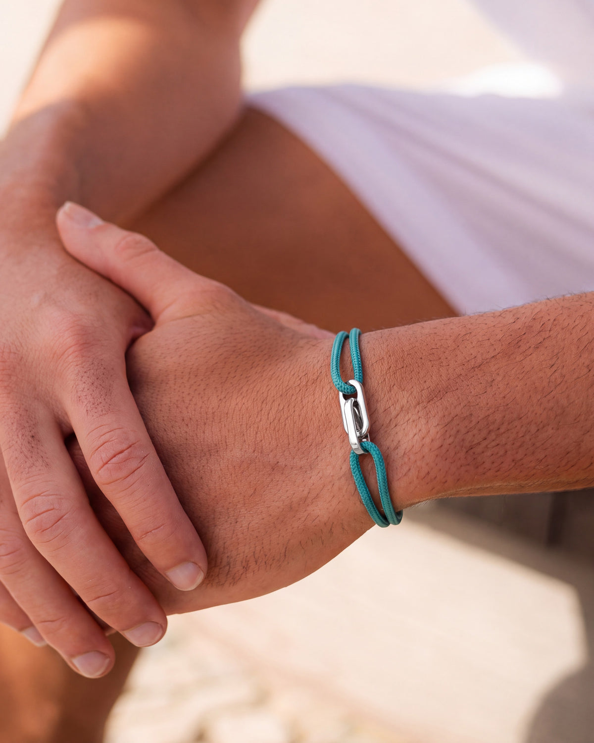 Atlantis silver bracelet with teal cord worn on the wrist, hands resting together in sunlight