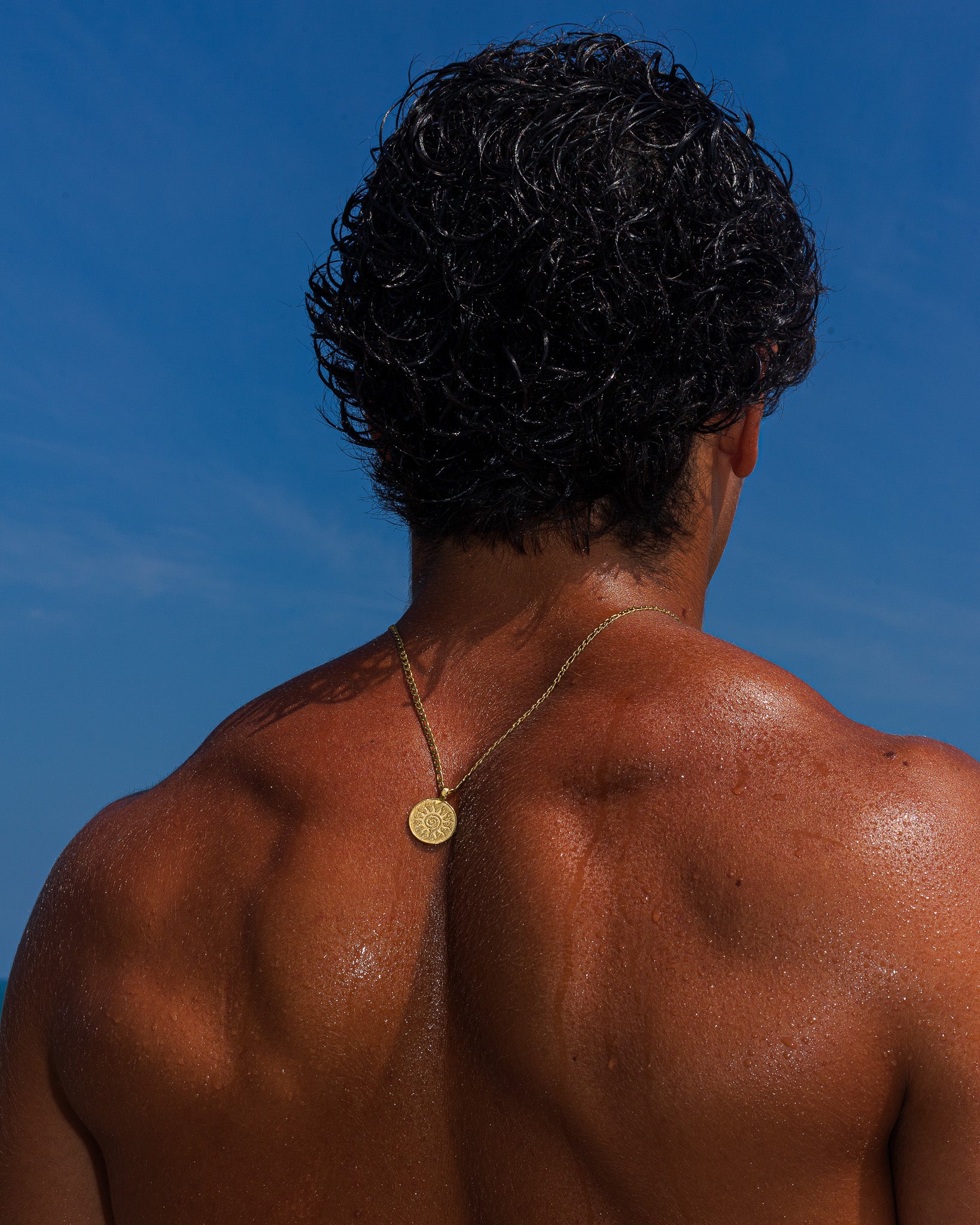 Back view of a person with wet dark hair wearing a thin gold chain necklace against a clear blue sky.