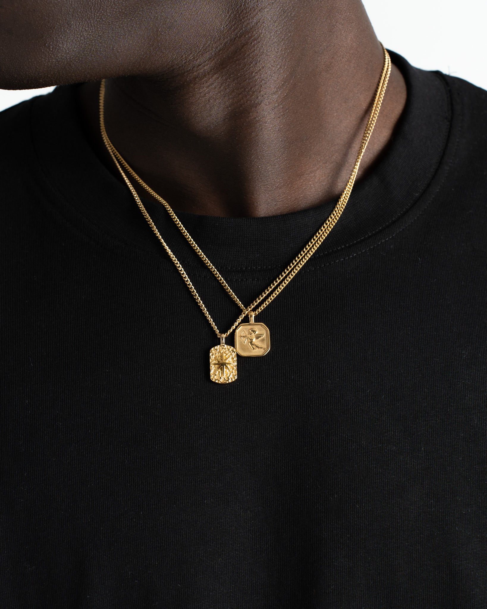 Gold chain necklace with two pendants a square engraved Cupid design and a textured rectangular charm on black fabric