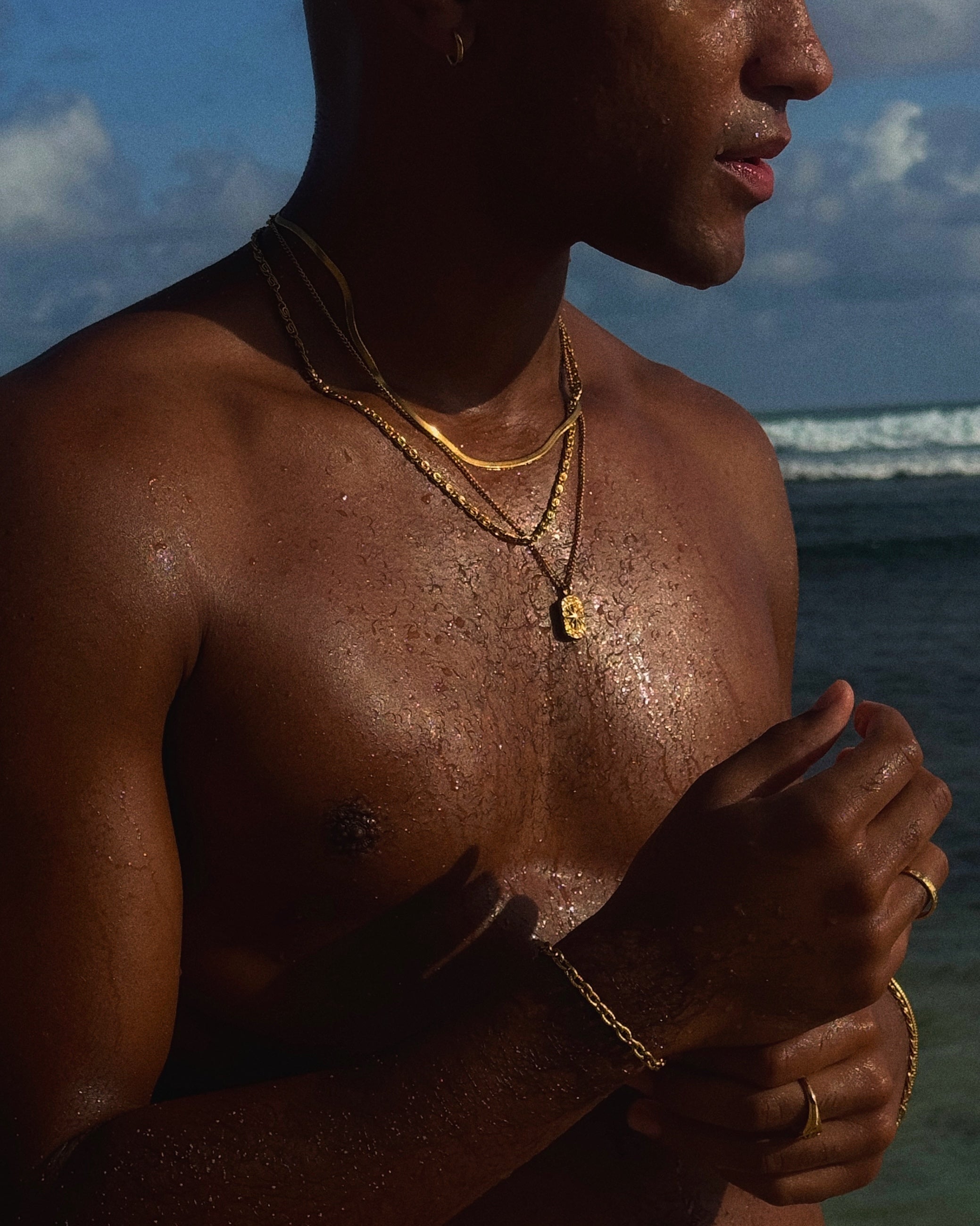 Layered gold chains around the chest with a square pendant outdoors by the sea