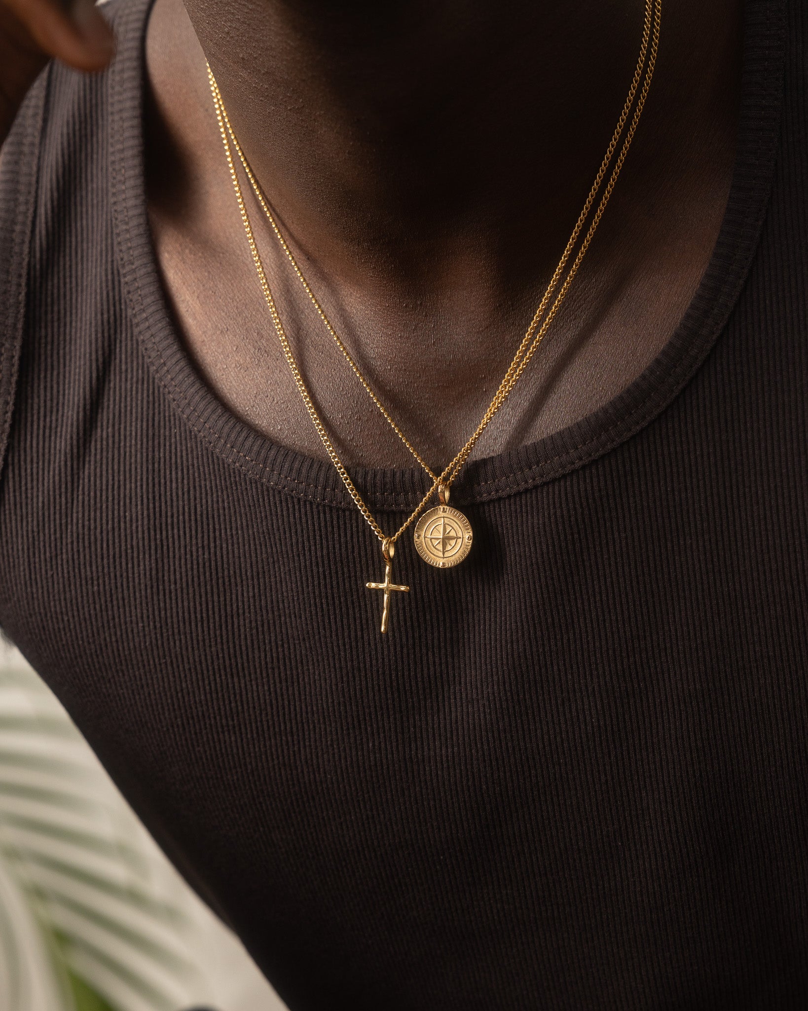 Man in a dark tank top wearing two layered gold chains with a cross pendant and a compass medallion pendant.