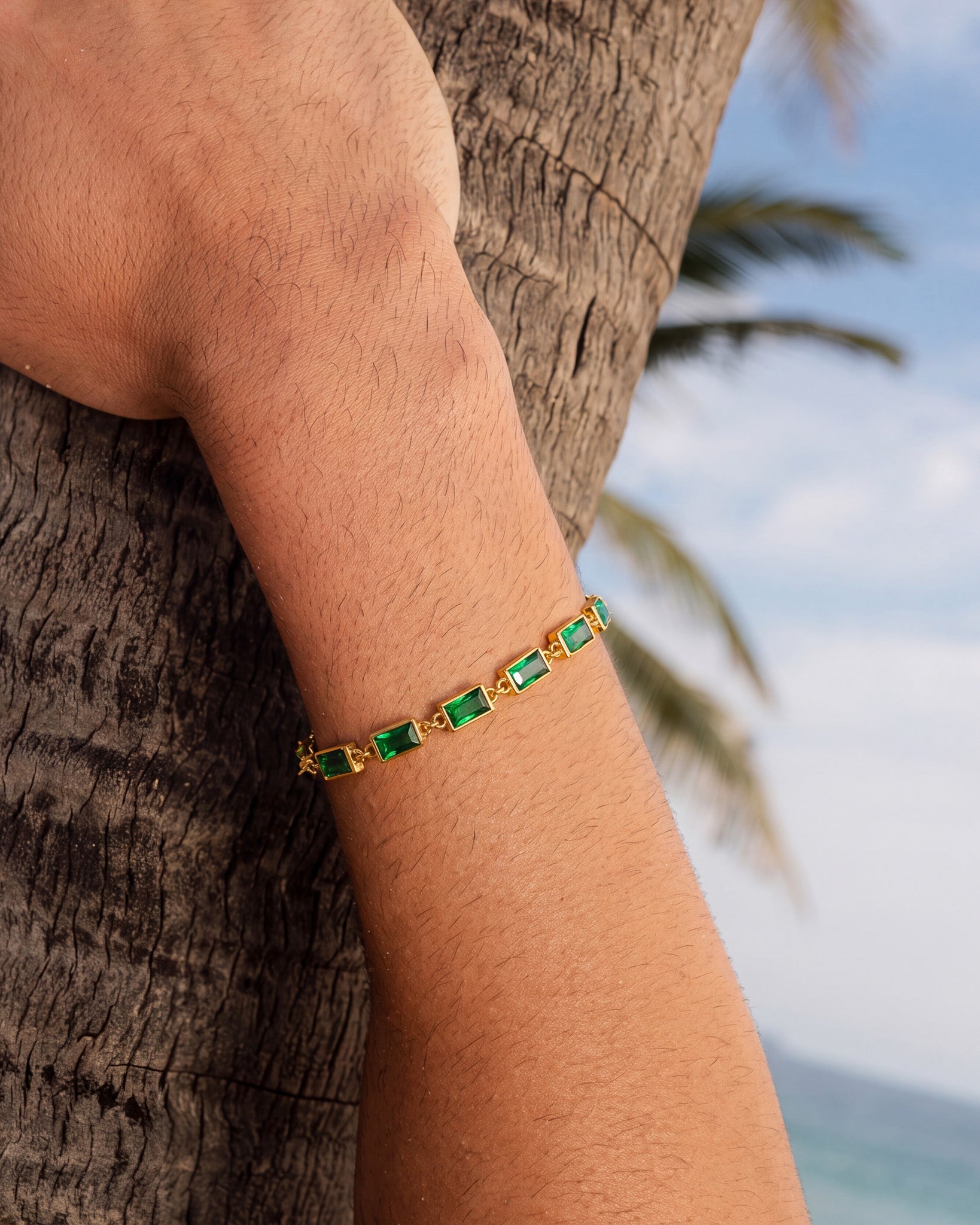 Close-up of the OMEN gold bracelet with emerald green stones on the wrist outdoors beside a tree trunk