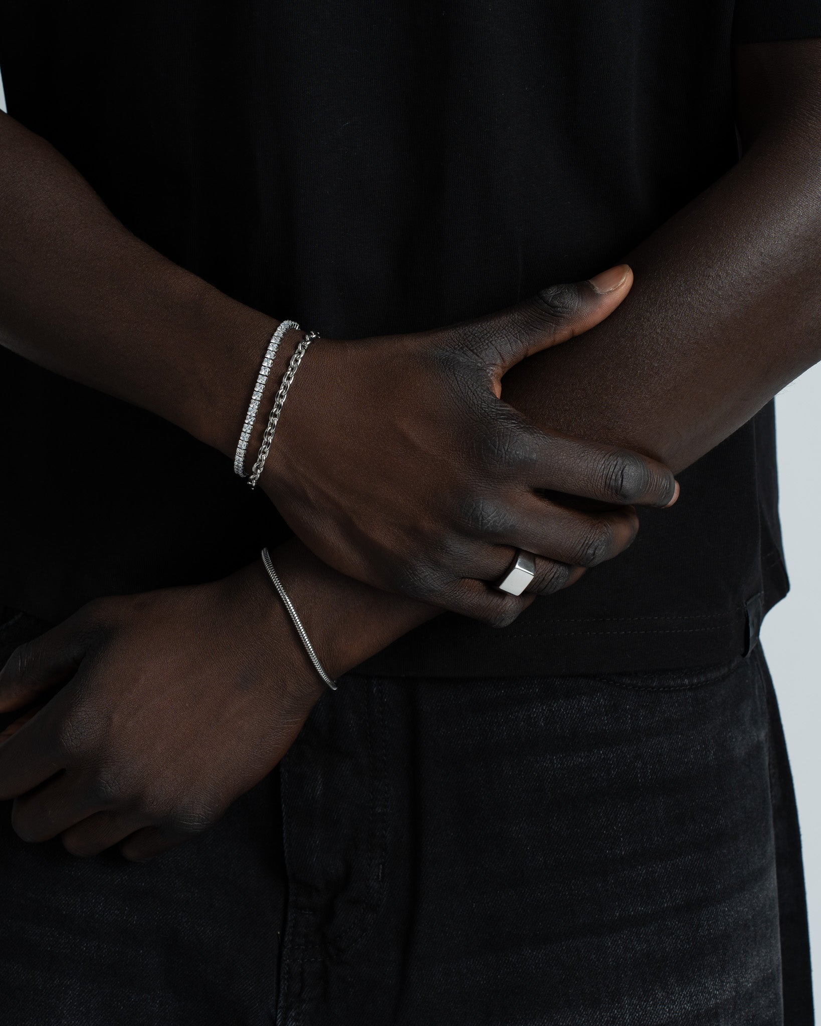 Dark-skinned forearm wearing two silver bracelets a diamond-studded band and a chain bracelet against a black shirt